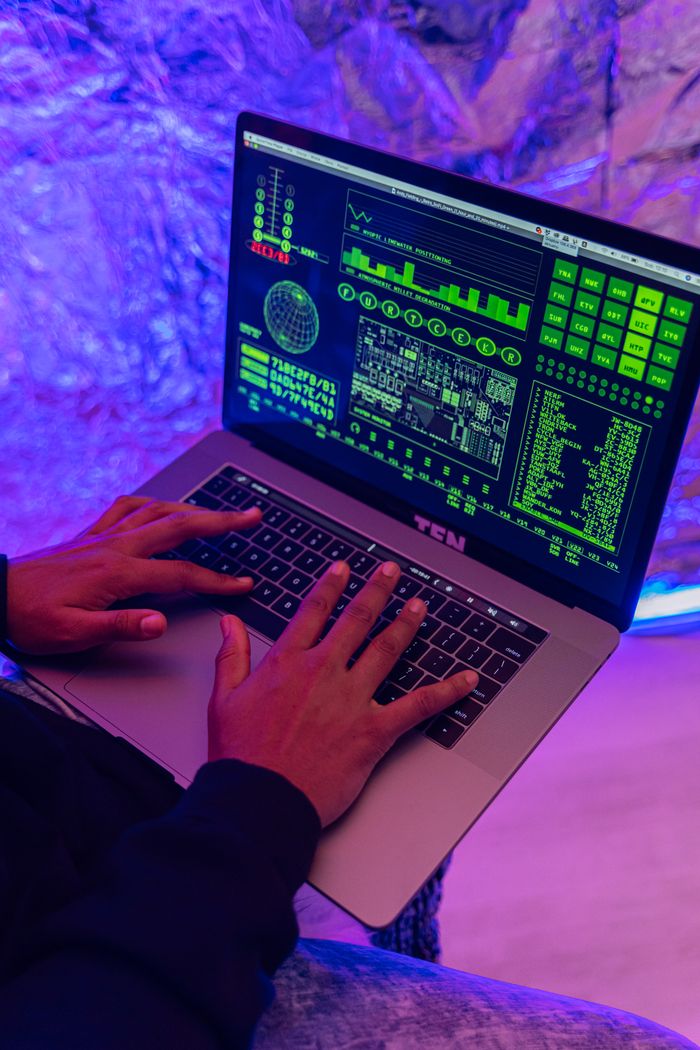 services-03 Close-up of hands typing on a laptop displaying green code, indoors, purple lighting.