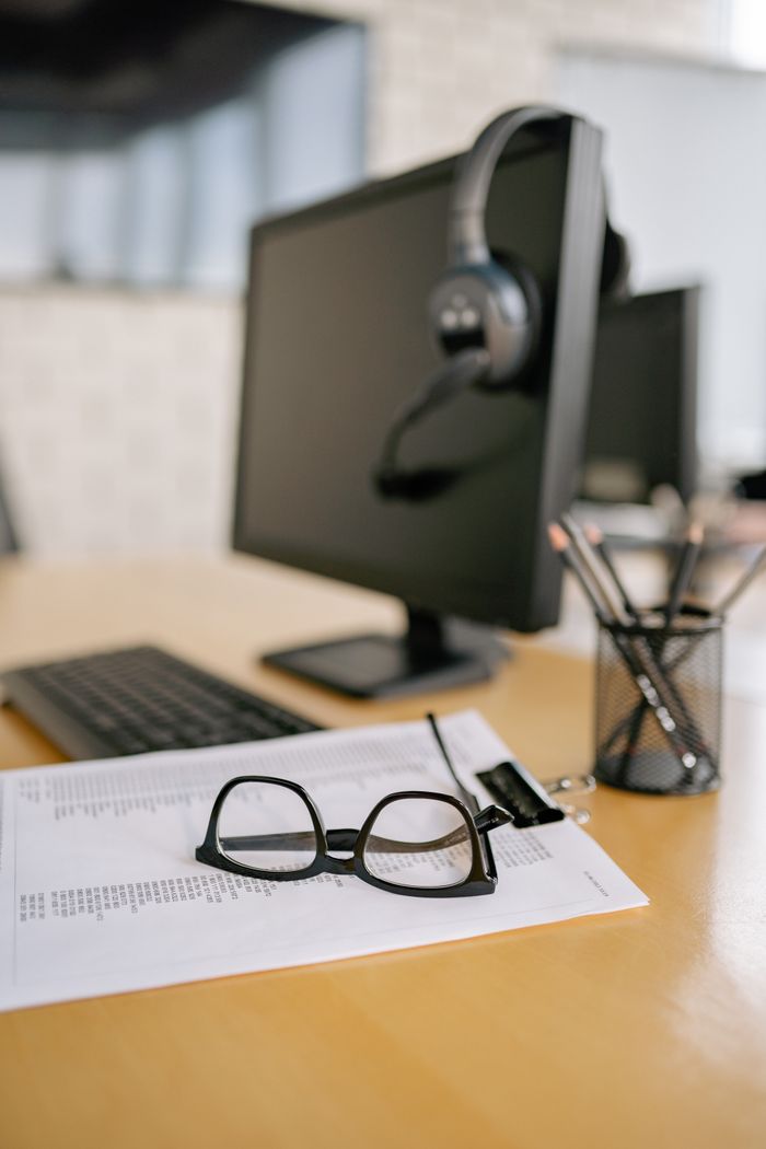 who-we-are Eyeglasses on a paper next to a computer and headset on a wooden desk.